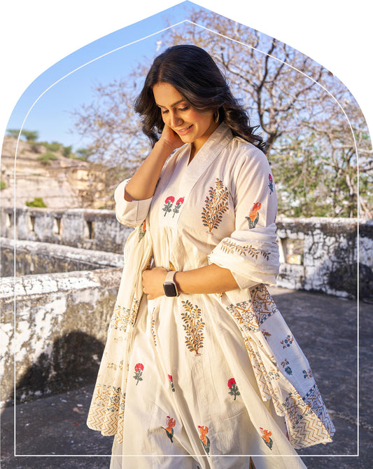 Woman in a white embroidered traditional outfit standing outdoors with stone architecture in the background. 